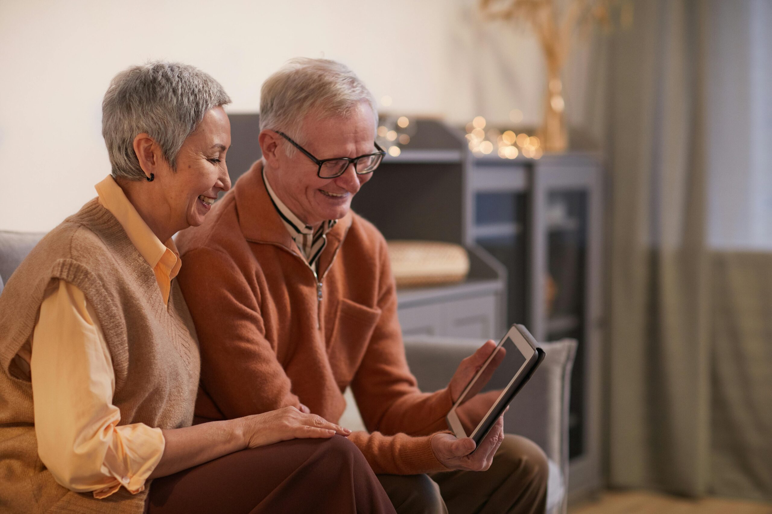 Two elderly individuals sitting on a couch, one holding a tablet, in a cozy living room setting with soft lighting and decorative elements in the background.