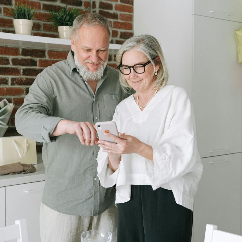 A caregiver in a modern kitchen guides a senior on using a smartphone, fostering independence and connection. The warm setting reflects compassionate in-home senior care, dignity, and personalized support.