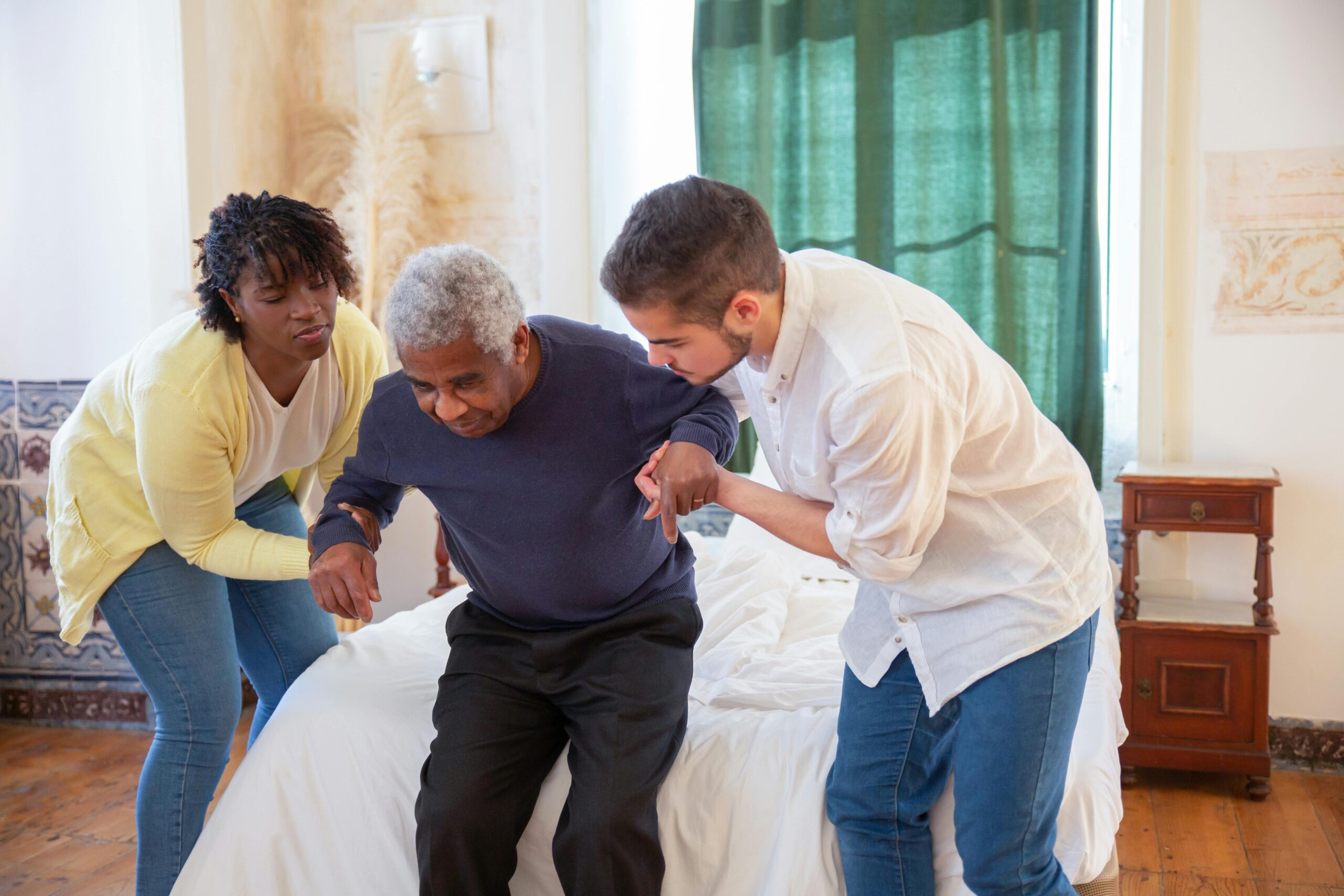 Two caregivers assisting a senior in standing up from a bed, demonstrating senior bathing assistance to prevent slips, falls, and discomfort at home.